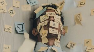A man's face is largely covered by post it notes on which are written buzzwords and symbols typical of the jargon of management