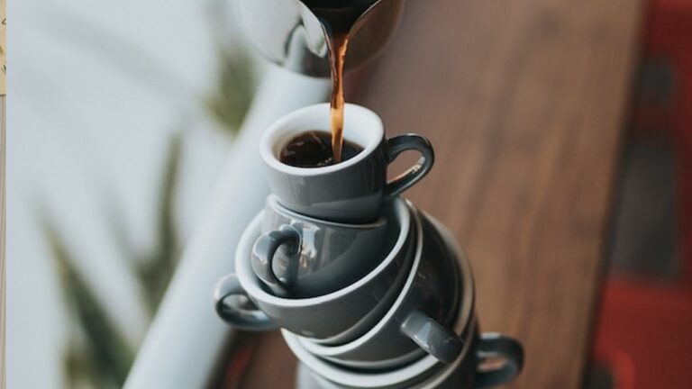 Black coffee is poured into a small cup which is propped precariously atop a tower of other coffee cups
