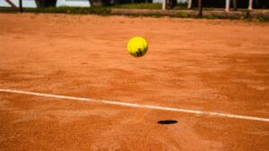 A yellow tennis ball in the air a few inches about a clay court, with one of the white lines in view