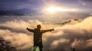a man standing on a mounatain top, hands outstretched, against a background of clouds