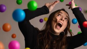 A close up on a woman in black polo neck panicking surrounded by bright coloured juggling balls