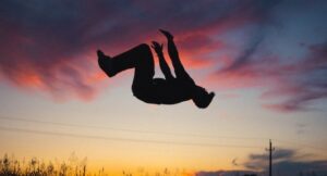 a silhouette of someone doing a back flip, midair, against a reddish evening sky with clouds