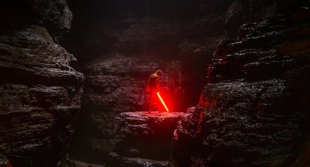 a jedi with his lightsabre, glowing red, allowed to point to the ground, at the centre of a dark, cavernous background