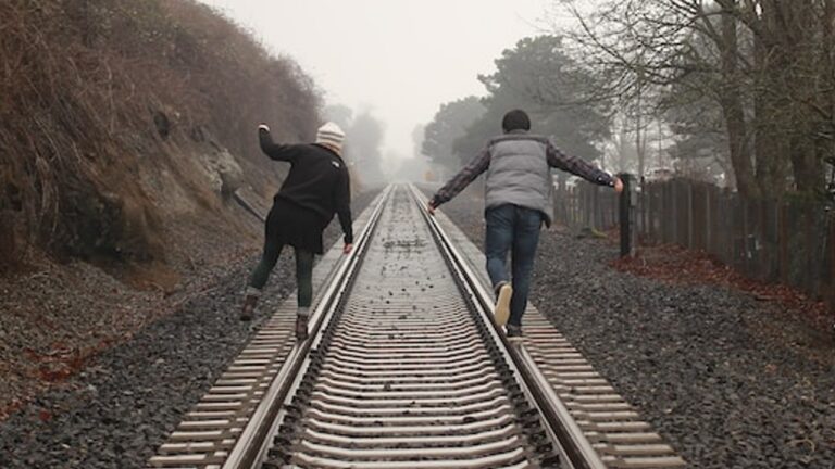 two figures balance on one foot, each standing on a different rail of a train track. They have their backs to us, and the track disappears into the distance