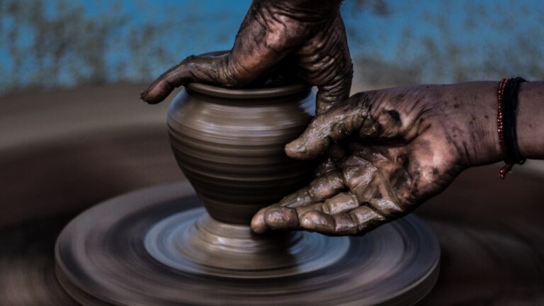 a pair of clay-covered human hands shaping a pot on a potter's wheel