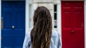 we see a woman with dark hair over her shoulders, in front of two doors, one blue, the other red