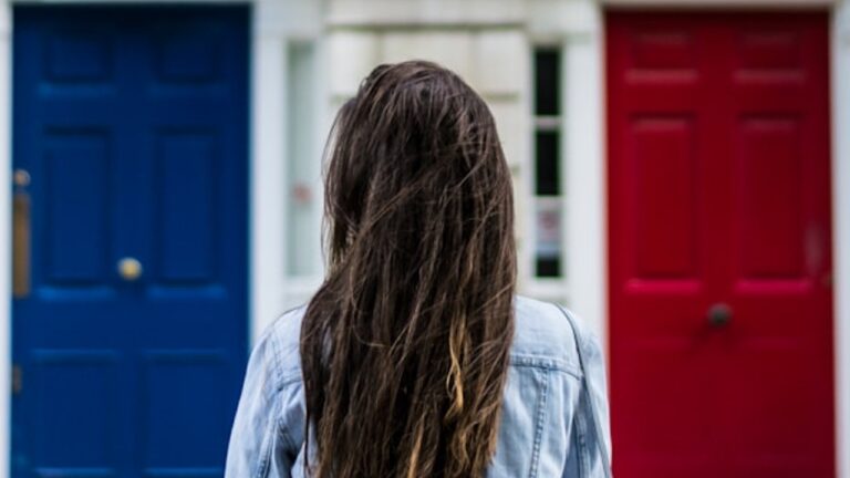 we see a woman with dark hair over her shoulders, in front of two doors, one blue, the other red