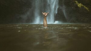 a hand signalling with two fingers thrust from a pool of water with a waterfall in the background